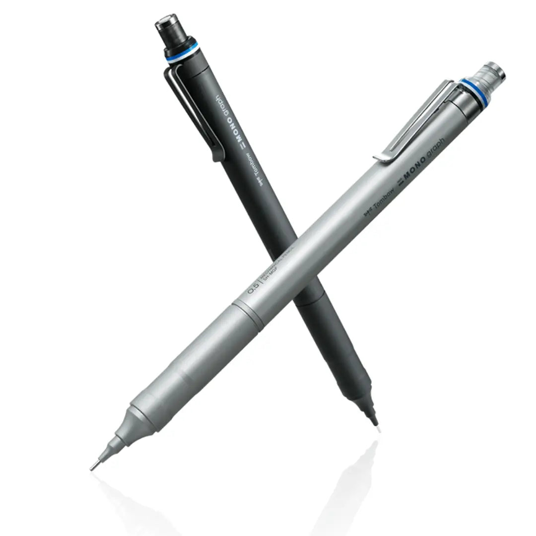 Tombow Mono Graph Fine siyah ve gümüş iki kalem çapraz duruyor, mavi halka detayı görünür