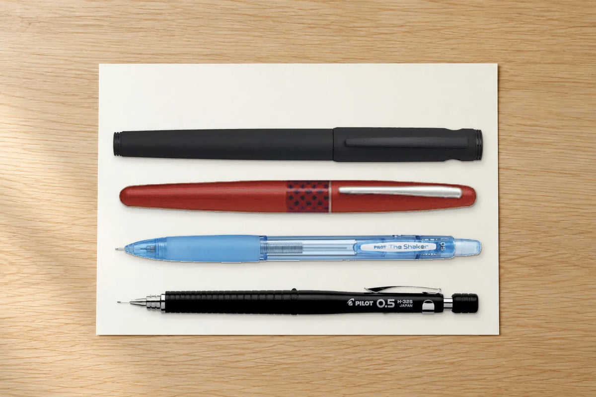 Meşe masada Pilot Explorer, kırmızı MR 3 Retro Pop, mavi The Shaker ve H32 kalemler, altında boş krem kâğıt