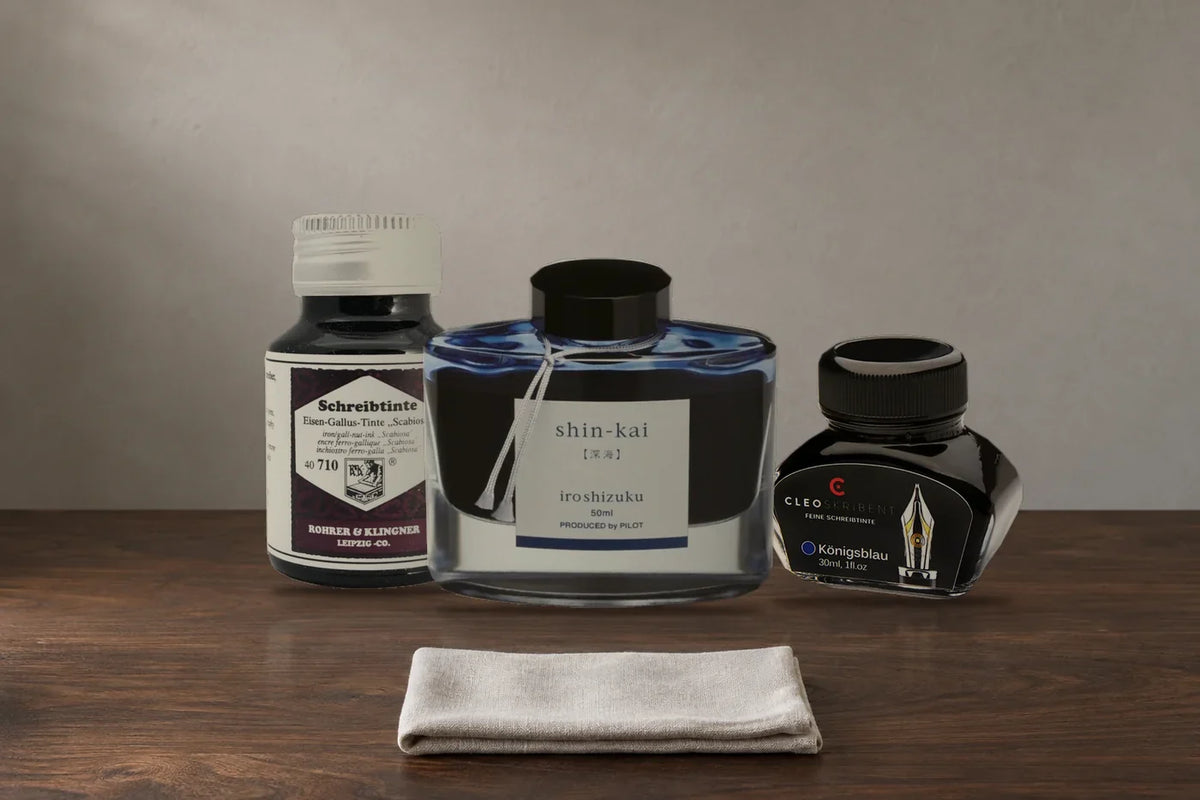 Koyu ahşap masada kapakları kapalı Rohrer & Klingner Scabiosa, Pilot Iroshizuku Shin-Kai ve Cleo Skribent Königsblau mürekkep şişeleri; önlerinde katlanmış keten bez.