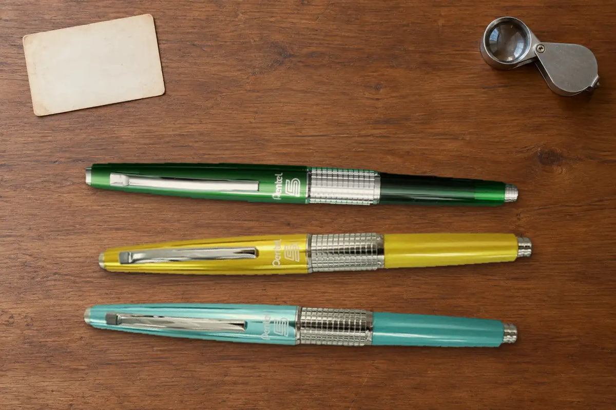 Ahşap masada yan yana Pentel Kerry Retro Green, sarı ve mavi versatil kalemler; yanlarında büyüteç ve boş bir kart