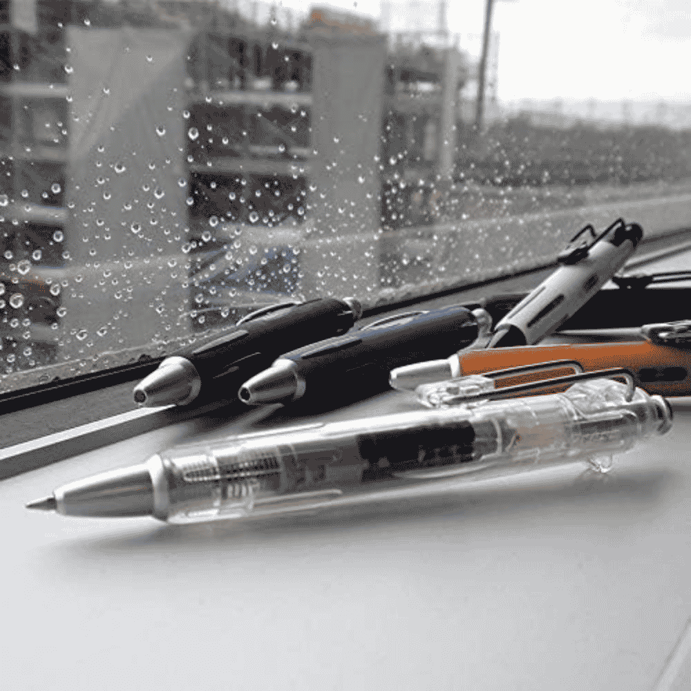 Tombow Airpress şeffaf kalem yağmurlu pencere kenarında, arkada siyah ve turuncu diğer modeller