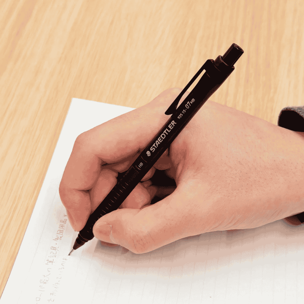 Staedtler 925-15 kalem elde tutulurken ahşap masada çizgili kağıda yazı yazılıyor