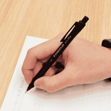 Staedtler 925-15 kalem elde tutulurken ahşap masada çizgili kağıda yazı yazılıyor