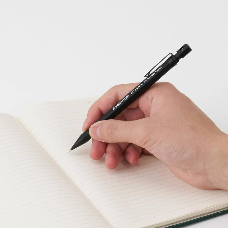 Staedtler Hexagonal kalemin siyah versiyonunu tutan el, çizgili açık defter üzerinde yazarken